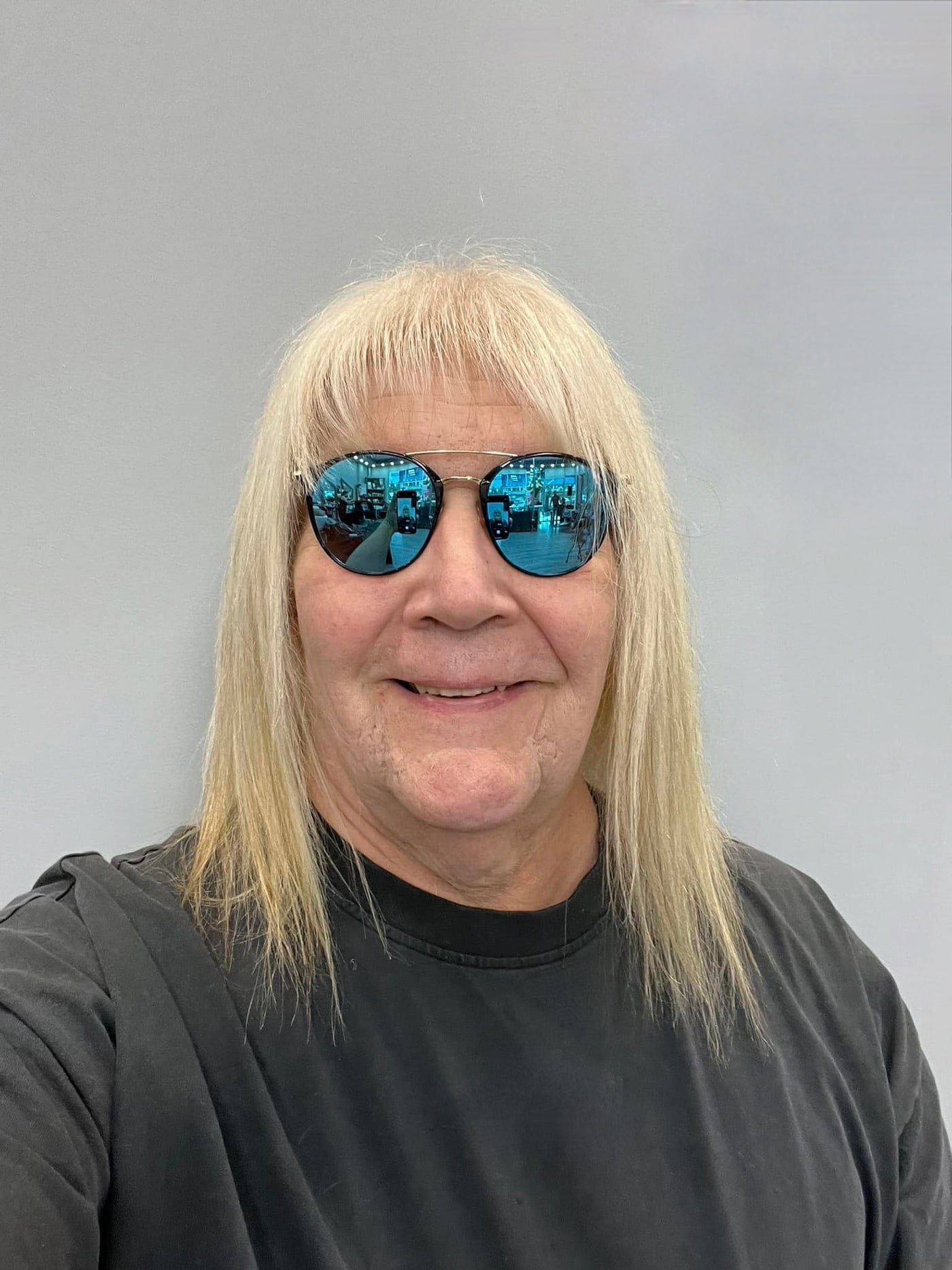 Portrait of Daniel Harris, smiling, wearing blue mirrored sunglasses and a dark t-shirt against a grey wall.
